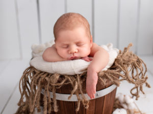 This is a newborn photography image of newborn baby in brown bucket with white backdrop