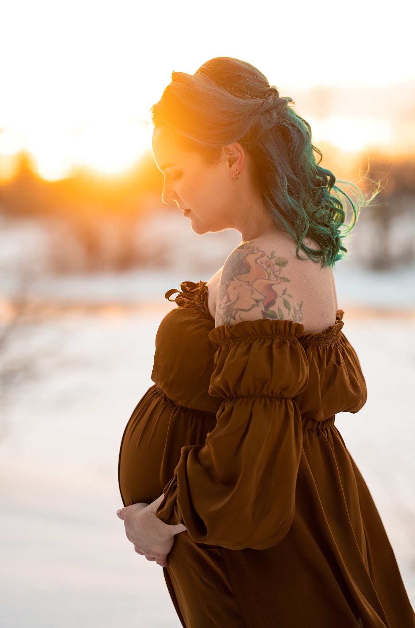 Mother to be looks down at her bump in a snowy field at sunset