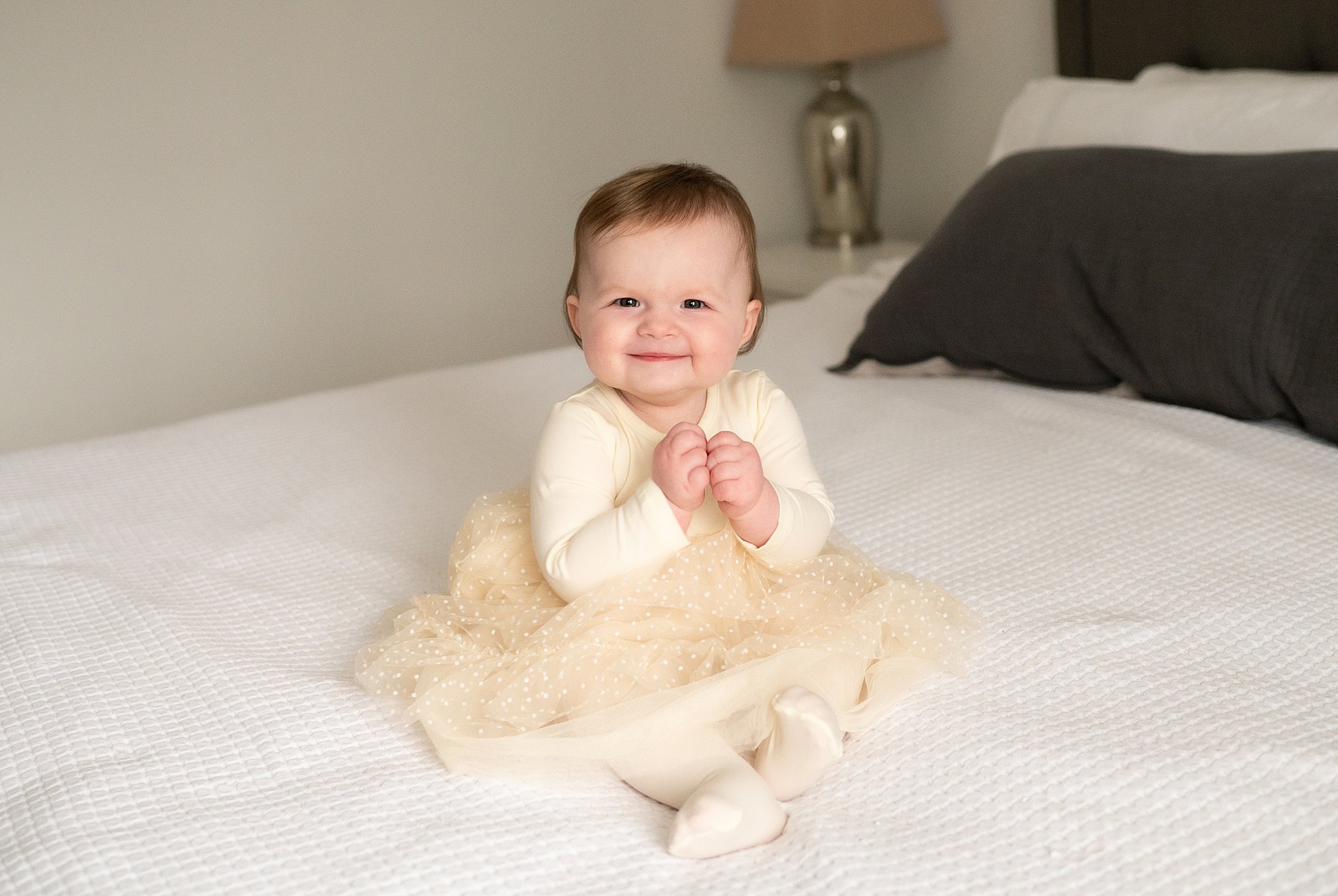 A toddler sits on a white bed in a yellow dress with their hands together and a big smile