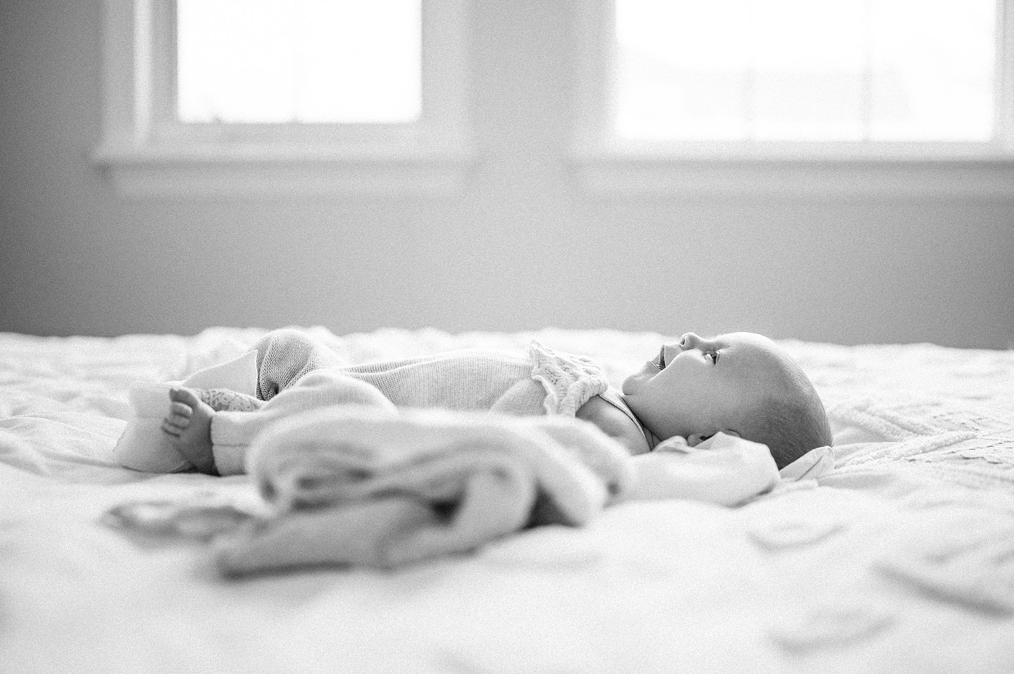 A newborn baby lays on a large bed in front of windows in a onesie with a lace collar smiling baby enroute ottawa