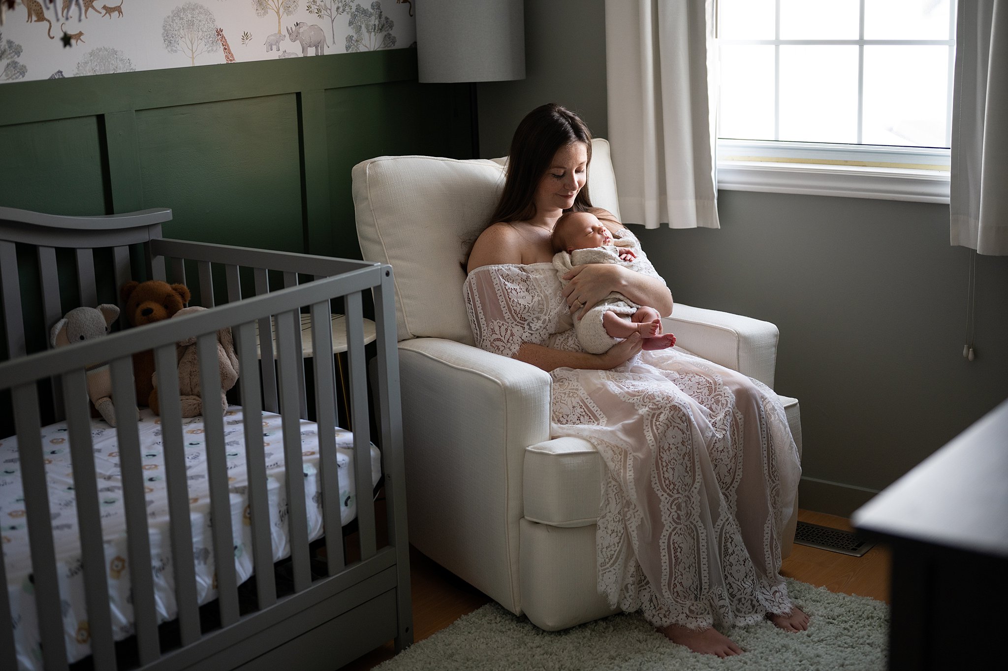 mother in a white lace dress sits in a nursery chair holding newborn under a window