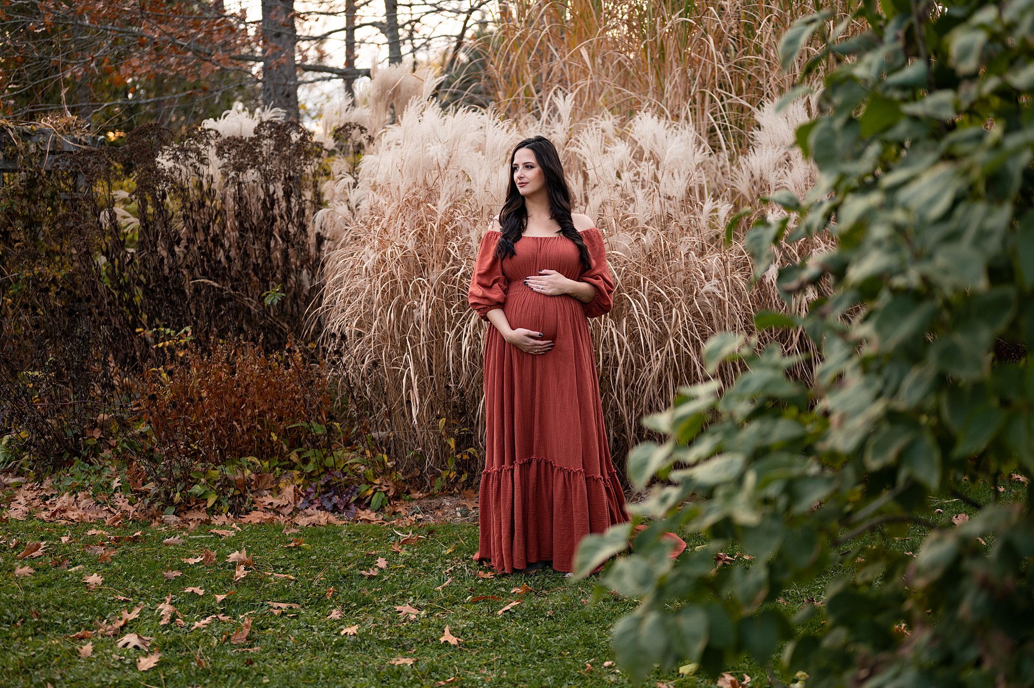Mother to be stands amongst tall grasses in a maternity gown while holding her bump ottawa midwives