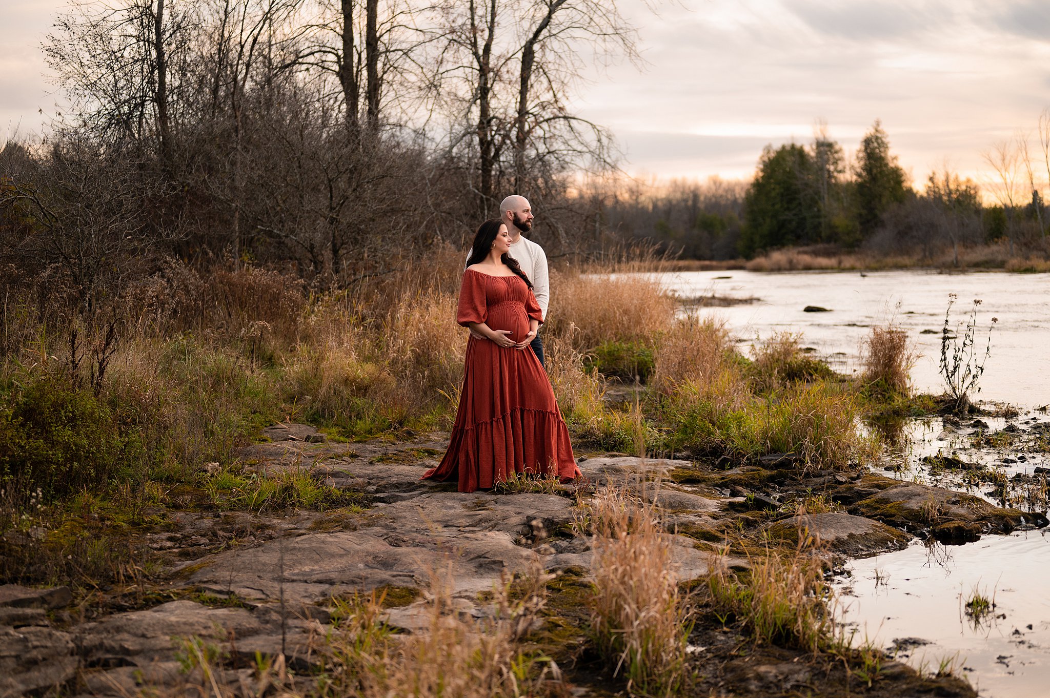 A couple stand on the edge of a river in the woods while holding soon to be mother's bump ottawa midwives