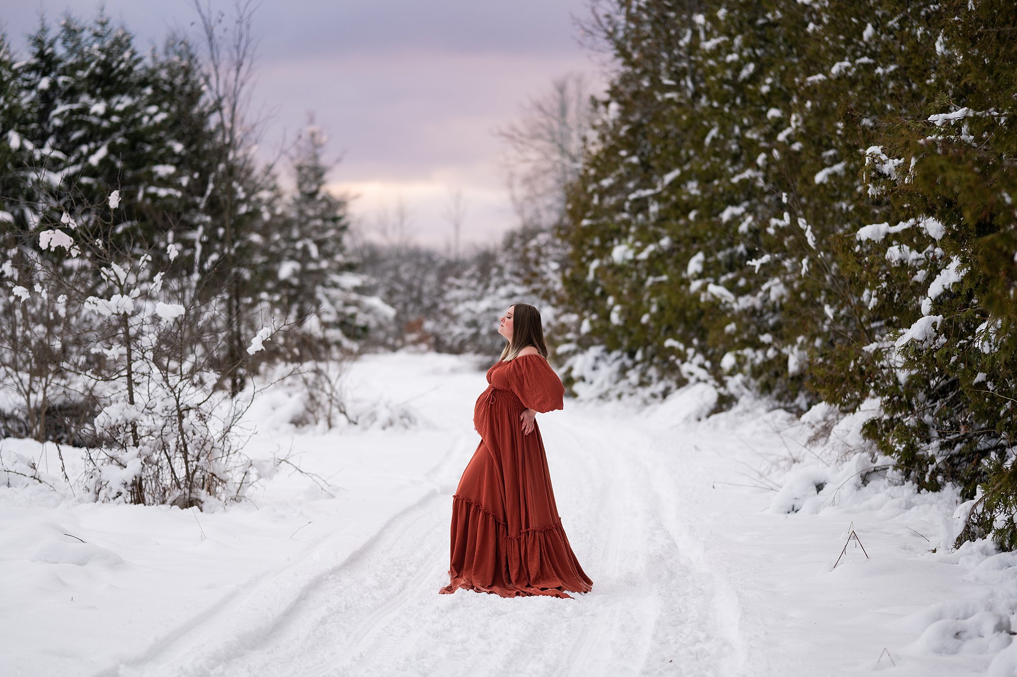 Mother to be stands in a maternity gown in a snowy path in the woods ottawa midwives
