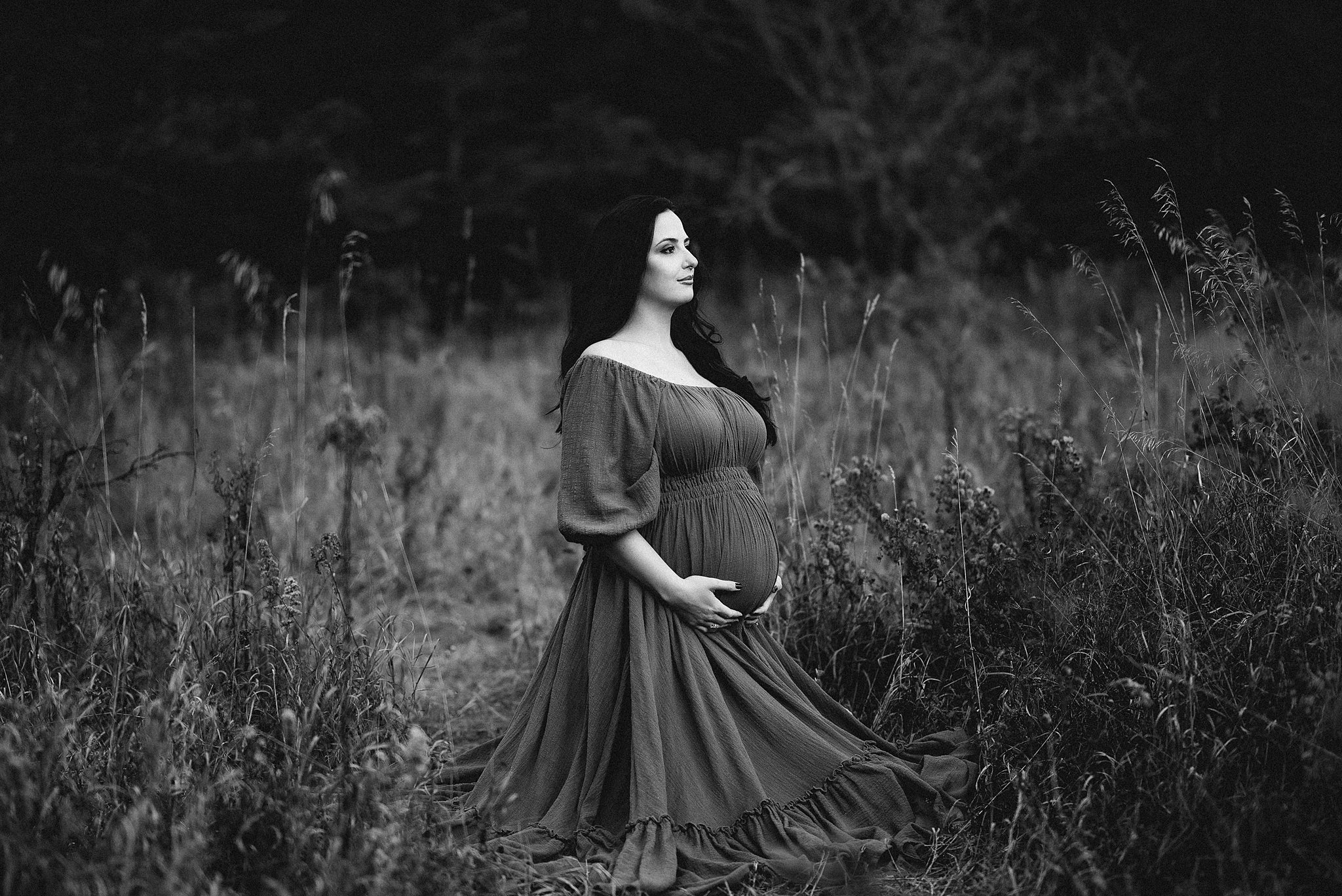 A mother to be kneels in a grassy field in a dark maternity dress surrounded by tall grass hautemama