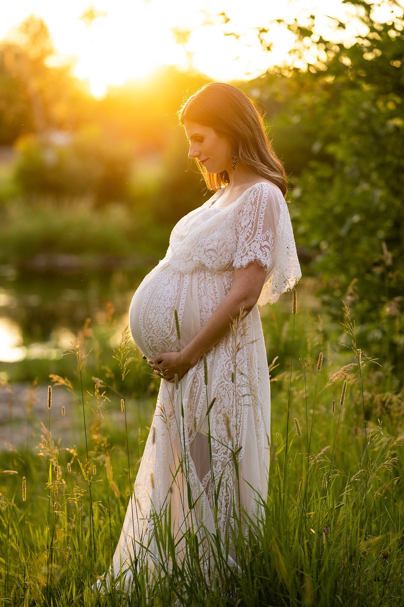 A mother to be stands in some tall green grass at sunset while looking down at her bump hautemama