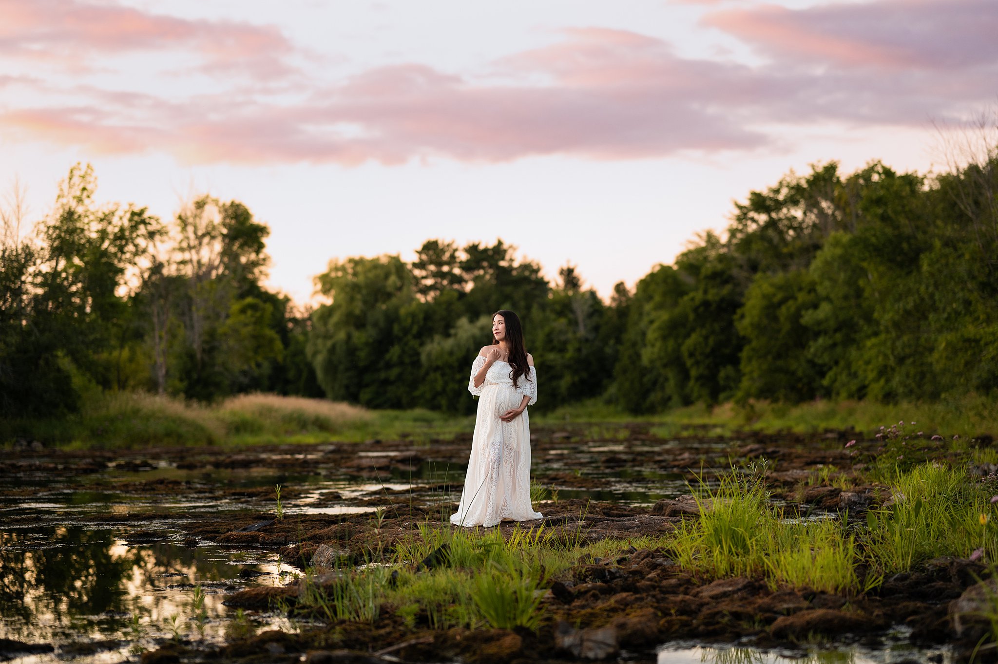 A mother-to-be stands in a white dress and holding her bump in a wetland with tall grass surrounded by trees hautemama