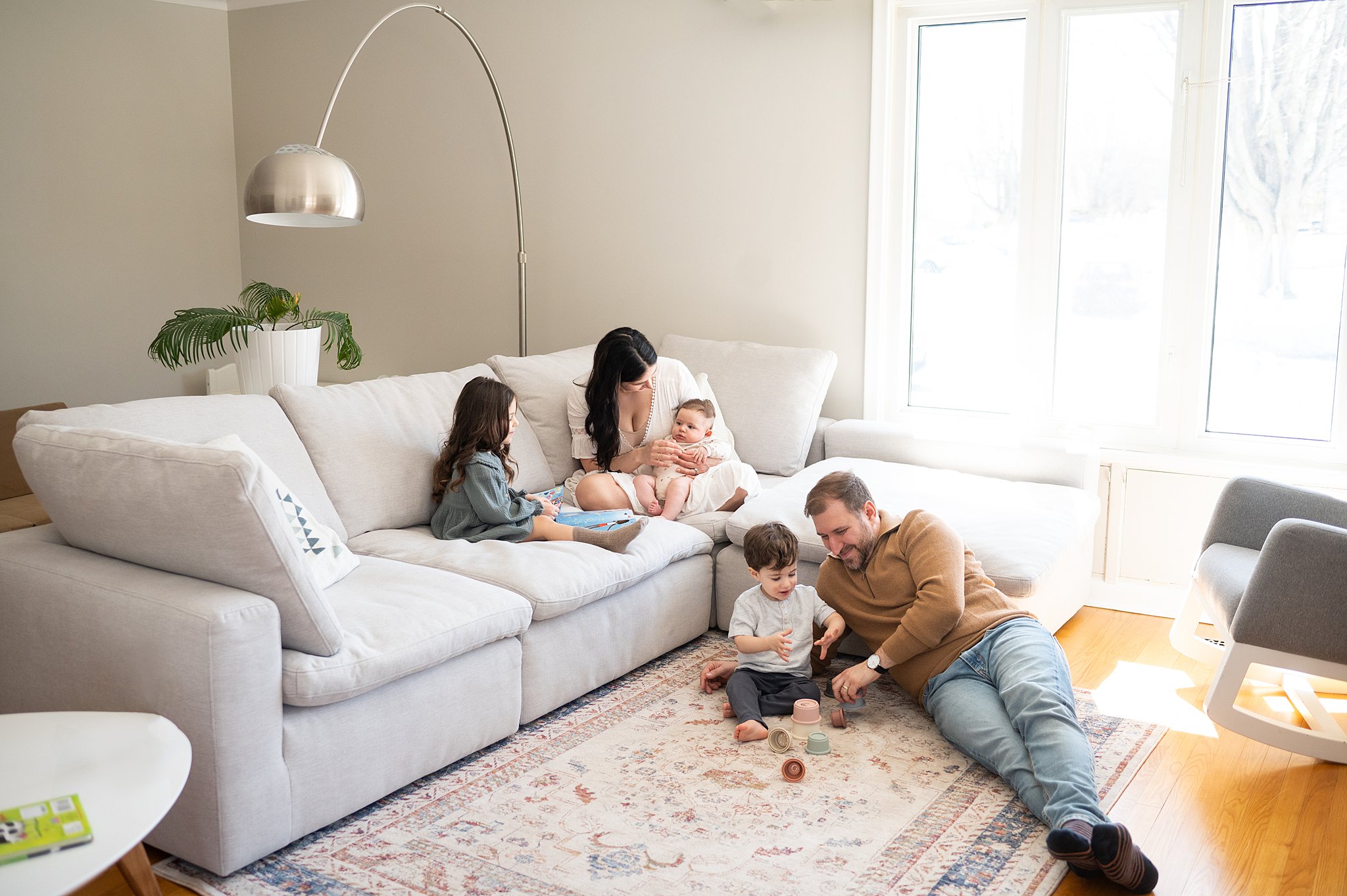 A mother sits on a couch playing with her newborn baby and toddler daughter while dad and another toddler boy play with some toys on the floor in front of them ottawa lactation consultants
