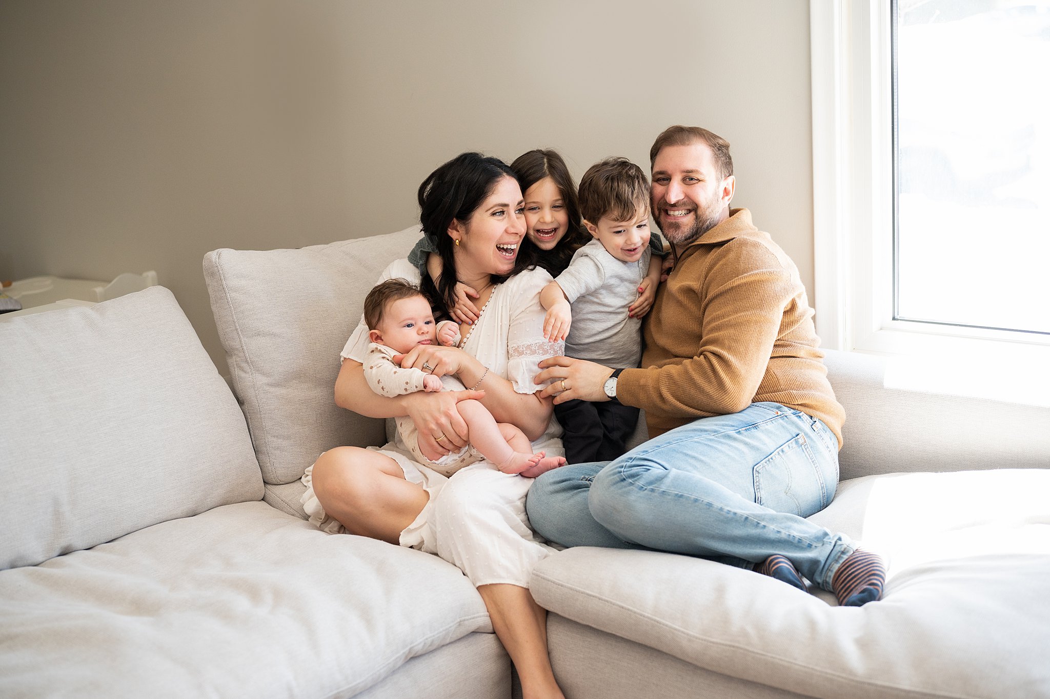 A family of five play in a corner of a large couch next to a window with mom holding the newborn baby ottawa lactation consultants