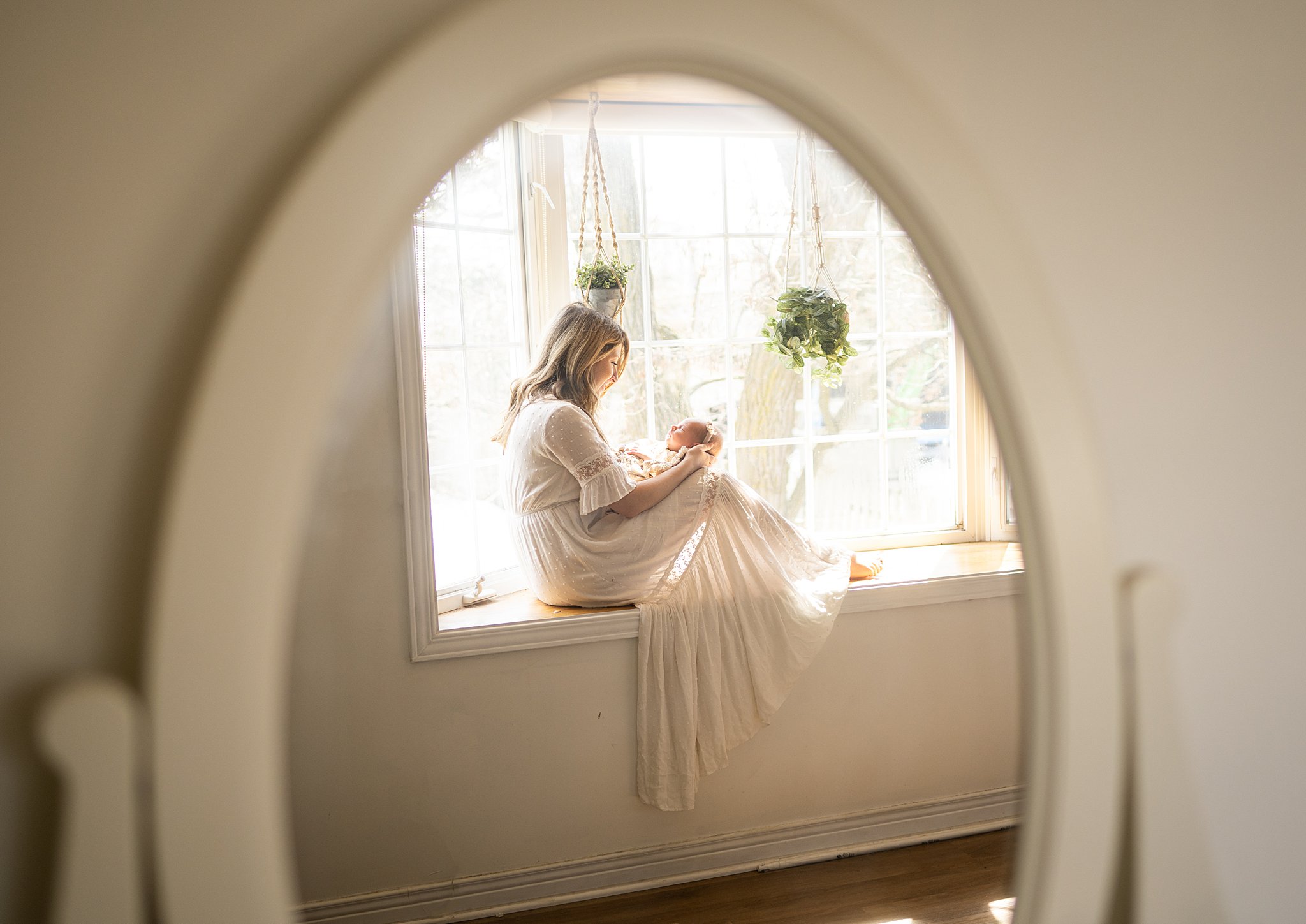 A mother in a white dress sits in a bay window looking down at her newborn baby in her lap ottawa prenatal massage