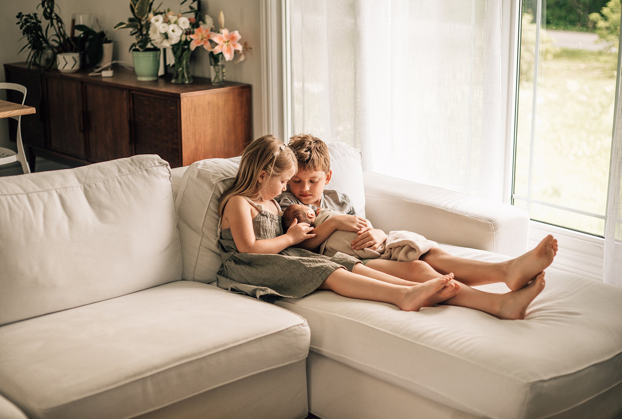 A brother and sister sit on a couch together while holding and cuddling their newborn sibling