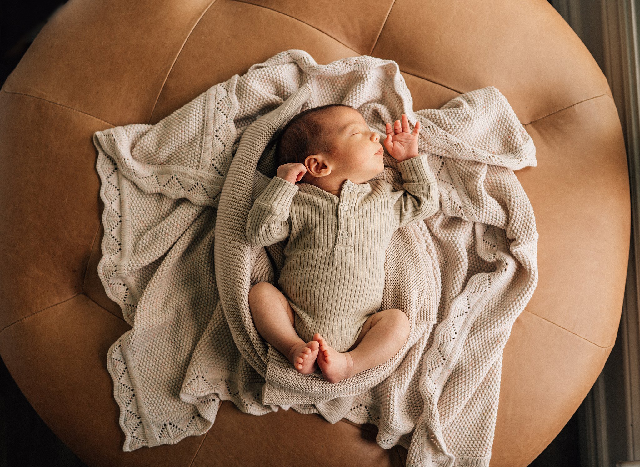 A newborn baby sleeps in a beige onesie on a tan leather ottoman with blankets ottawa prenatal yoga