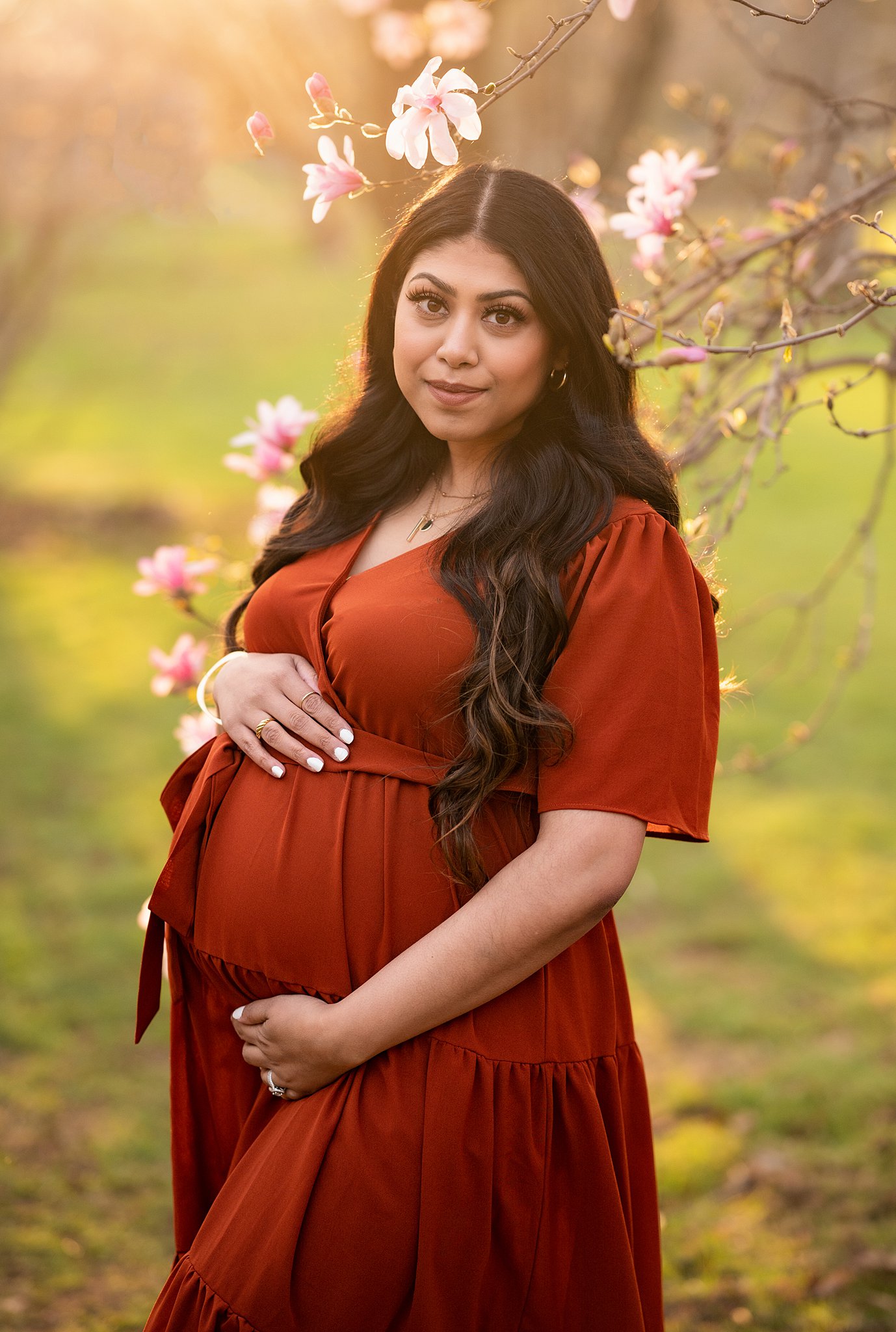 A mother to be holds her bump while standing under a tree blooming pink flowers