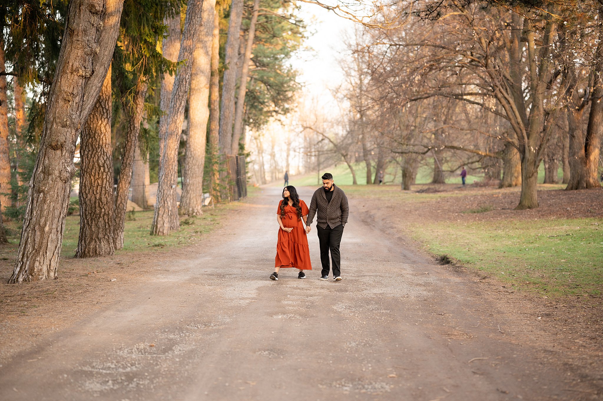 A mother-to-be in a red maternity dress walks down a park path lined with tall trees while holding hands with her husband ottawa 3d ultrasound clinics