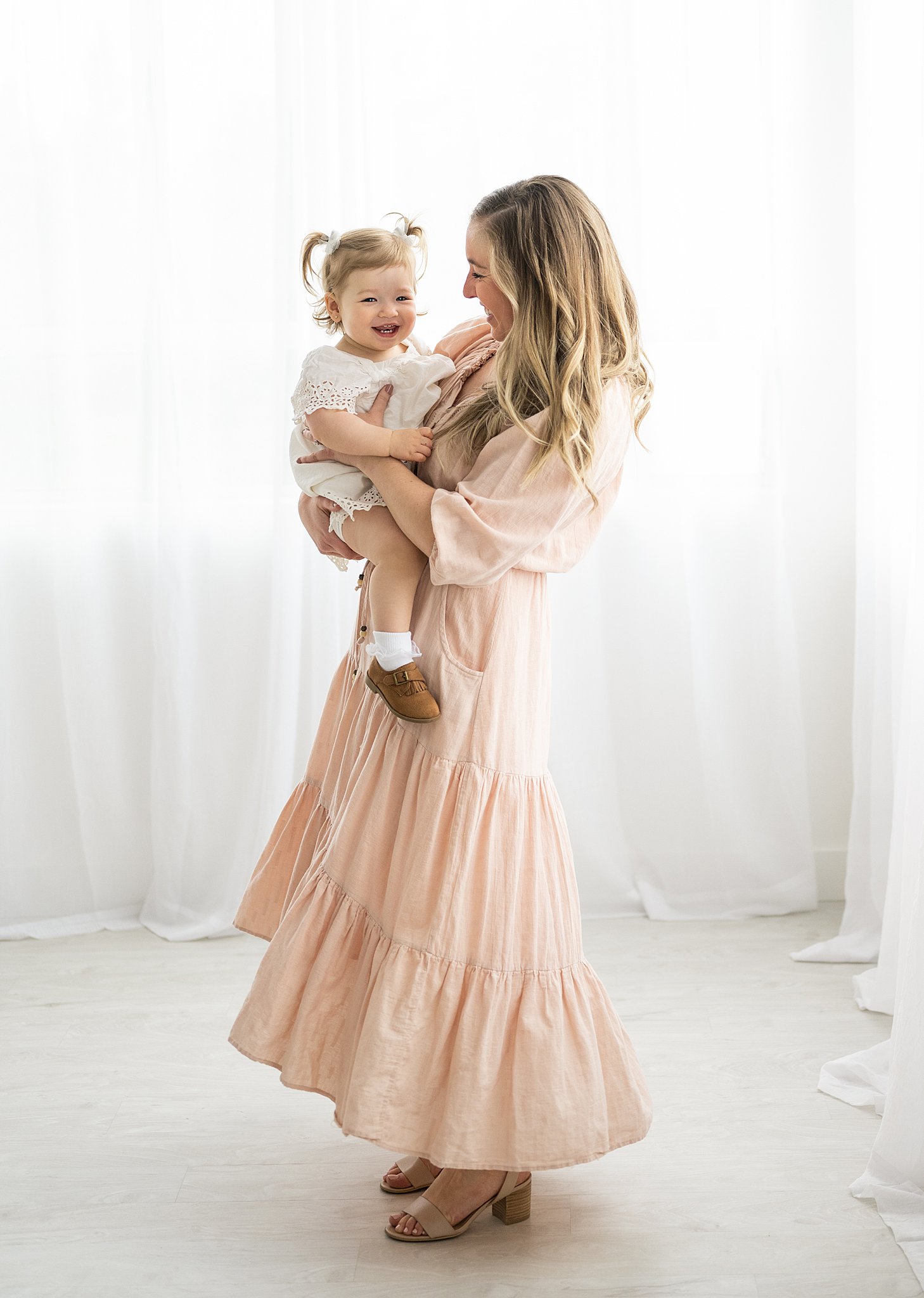 A mother in a pink dress stands in a studio bouncing her toddler daughter on her hip