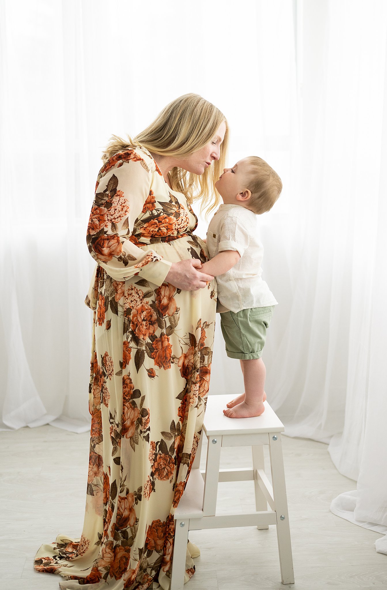 A pregnant mother in a floral dress leans down to kiss her toddler who is standing on a stool in a studio ottawa prenatal classes