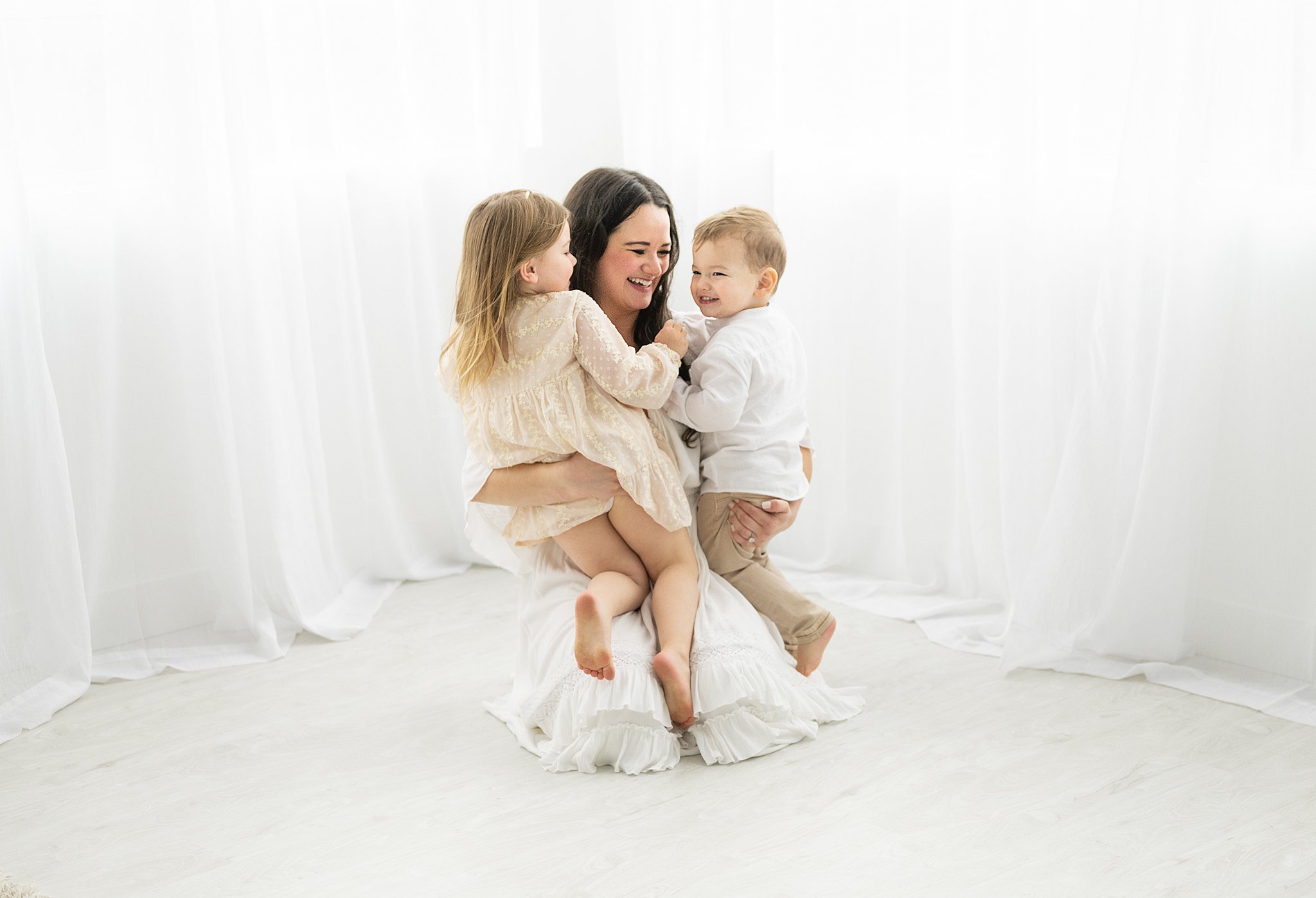 A mother in a white dress plays with her two toddler children while kneeling on the floor of a studio ottawa prenatal classes