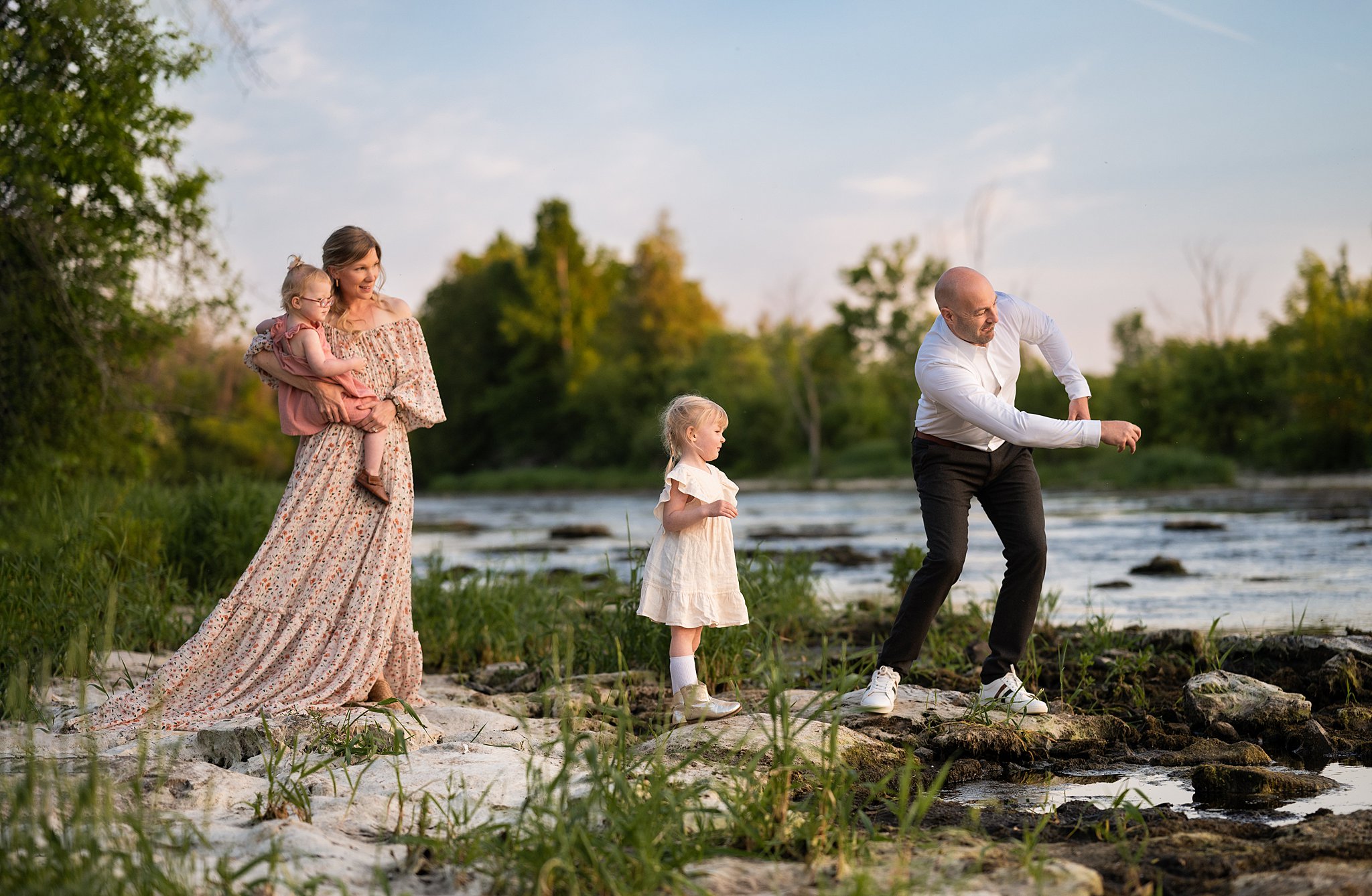 A father skips rocks across a river while mom and their two daughters look on Ottawa Nannies
