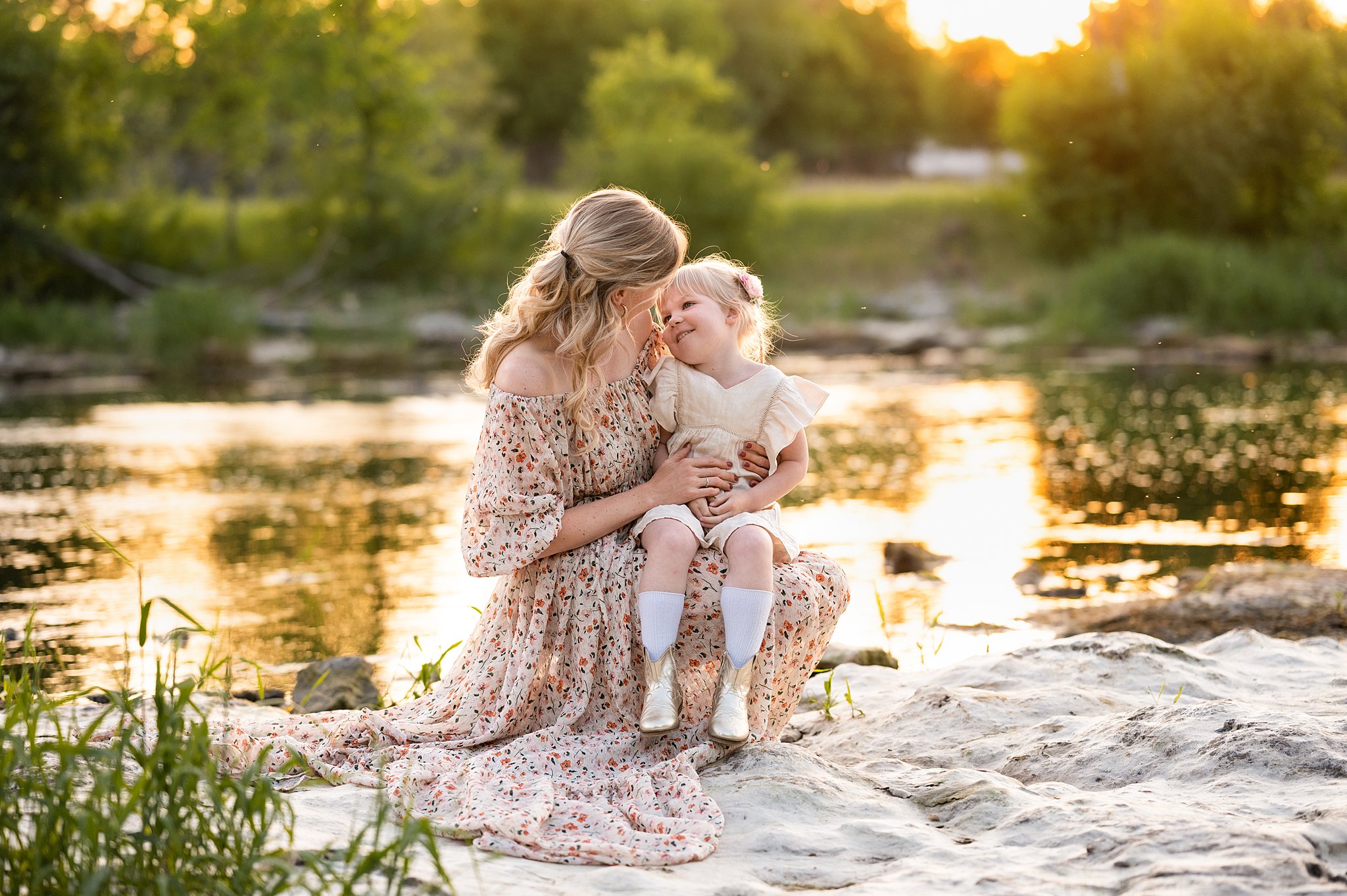 A mother in a long floral dress squats on a rock by a river with her young daughter in her lap Ottawa Nannies