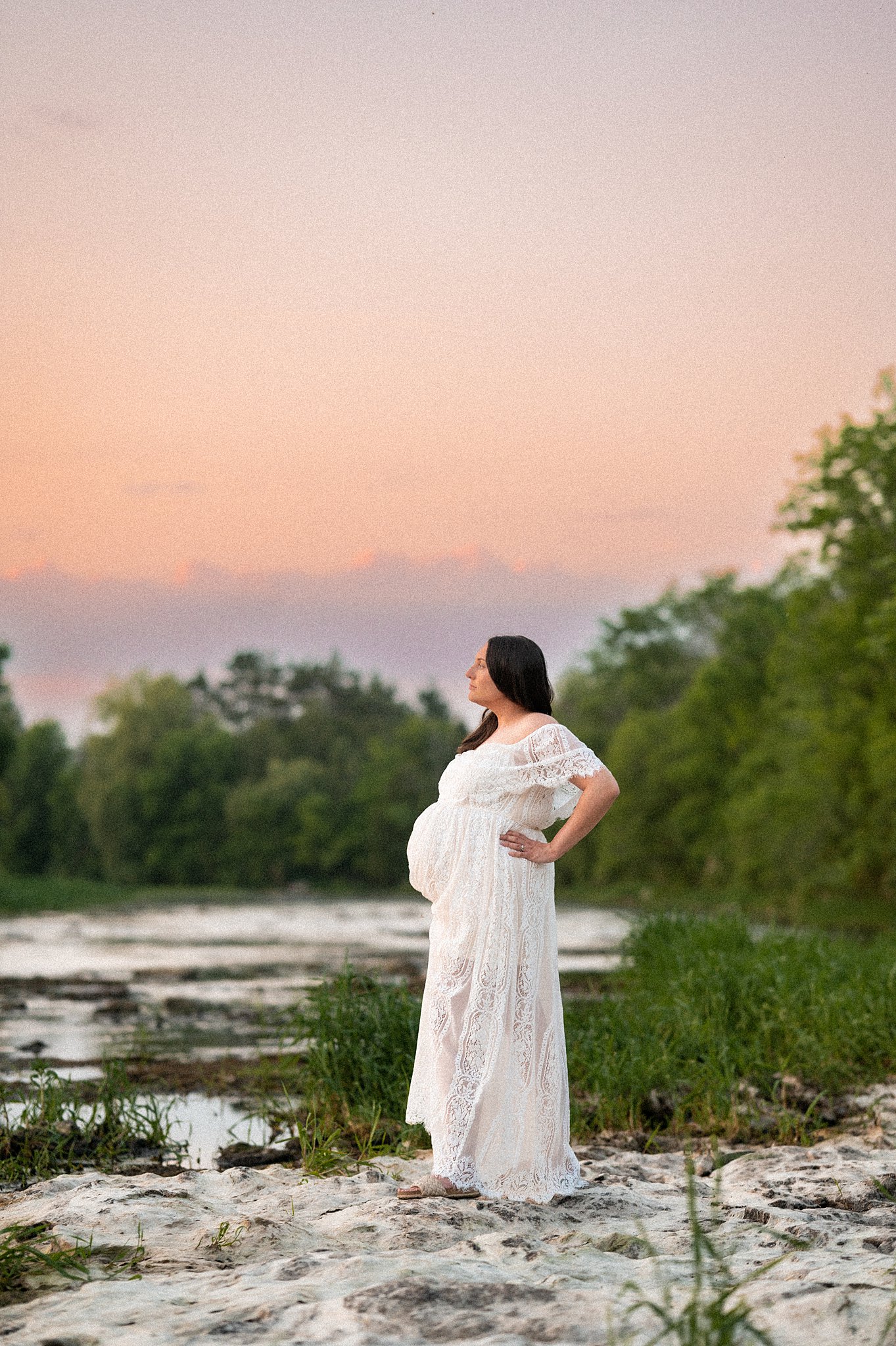 A mother to be in a white lace maternity gown stands on the edge of a river at sunset holding her bump night nurse ottawa